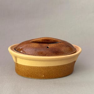 Op de afbeelding: Een ovale keramische ovenschaal met een tweekleurig ontwerp, bestaande uit een gespikkelde bruine basis en een gladde crèmekleurige rand. De schaal is voorzien van een bijpassend ovaal deksel met een donkerbruine glazuurlaag en een stoomgaatje.