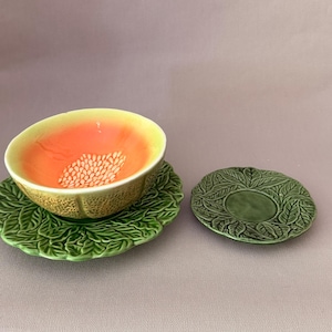 Op de afbeelding: Een keramische kom met een geel en oranje interieur met een wit zaadpatroon, rustend op een groen bladvormig schoteltje. Daarnaast ligt een bijpassend groen bladvormig schoteltje met een gladde cirkelvormige kern en gedetailleerde bladstructuur.