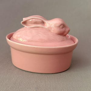 Może przedstawiać: Jasnoróżowe ceramiczne pudełko na drobiazgi ze zdejmowaną pokrywką. Pokrywka przedstawia wyrzeźbioną figurkę odpoczywającego królika z detalami uszu i fakturą futra. Podstawa pojemnika ma owalny kształt z gładkimi ściankami i błyszczącym wykończeniem.