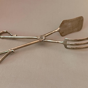Op de afbeelding: Een zilverkleurige metalen serveertang met een scharnierend ontwerp en een centraal rond draaipunt. Het ene uiteinde bestaat uit een massieve, rechthoekige platte spatel, terwijl het andere uiteinde een vork met drie tanden heeft.