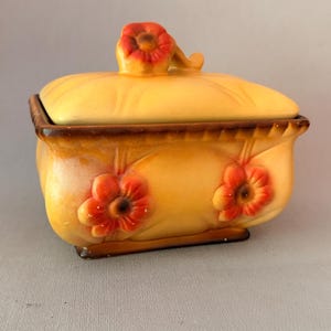 Op de afbeelding: Een rechthoekige keramische doos met deksel in een warme gele en oranje kleurverloop. Het object is voorzien van verhoogde, gevormde oranje bloemaccenten aan de voorzijde en een bijpassende bloemvormige handgreep op het deksel, met donkerbruine randen.