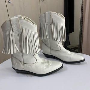 Könnte beinhalten: Ein Paar weiße Cowboystiefel aus Leder mit langen Fransen am oberen Schaft. Die Stiefel haben eine spitz zulaufende Zehenpartie, eine schwarze Sohle und den Markennamen Wrangler, der auf den Zuglaschen oben am Stiefelschaft eingeprägt ist.