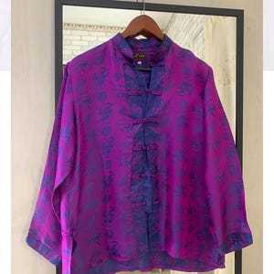 May include: A long-sleeved shirt in vibrant purple with a dark blue pattern featuring bamboo and circular motifs. The garment has a mandarin collar, a front opening secured by five fabric toggle closures, and a label reading "Lun" at the neck.