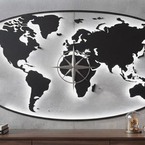 May include: An oval wall art piece featuring a black metal world map silhouette with a central compass rose. The map is backlit, creating a glowing white border against the wall. The design is contained within a thin black oval frame.