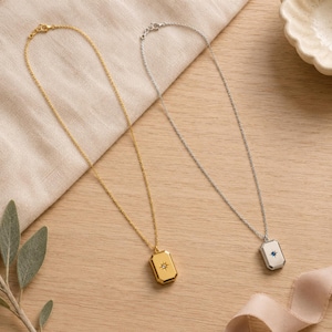 Op de afbeelding: Twee kettingen met rechthoekige hangers aan dunne kettinkjes. De ene ketting is goudkleurig met een heldere steen in een stervormig ontwerp. De andere is zilverkleurig met een kleine blauwe steen in een stervormig ontwerp op de hanger.