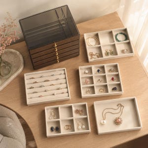 May include: A dark wood jewelry organizer with five drawers and a translucent dark gray lid, accompanied by four beige velvet-lined trays containing various rings, earrings, necklaces, and bracelets arranged in individual compartments.