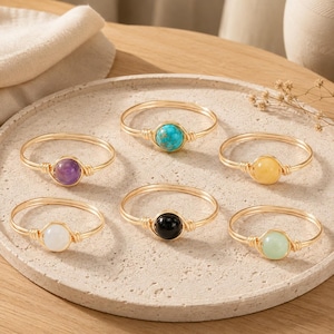 Puede incluir: Seis anillos de alambre enrollado con cuentas de piedras preciosas redondas en varios colores, incluyendo turquesa, morado, amarillo, negro, blanco y verde claro. Cada anillo está hecho de un alambre fino dorado alrededor de una piedra.