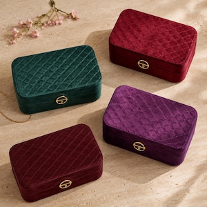 May include: Four rectangular jewelry boxes with a quilted velvet exterior in dark green, deep red, and purple. Each box features a gold-toned metal latch on the front. The lids have a diamond-stitched pattern, and the boxes are arranged on a surface.
