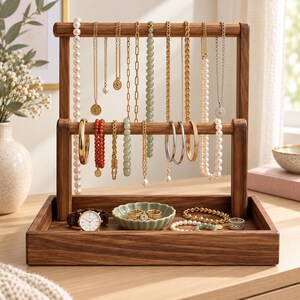 May include: A two-tiered wooden jewelry stand with a rectangular base tray. The stand features multiple hanging necklaces, including pearl, gold chain, and beaded styles. The base tray holds a wristwatch, a small scalloped dish, and bracelets.