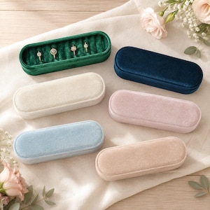 Puede incluir: Seis joyeros ovalados alargados con forro de terciopelo y tapas a juego. La colección incluye colores verde esmeralda, azul marino, crema, rosa empolvado, azul claro y beige. La caja verde está abierta y muestra cuatro anillos en su interior.