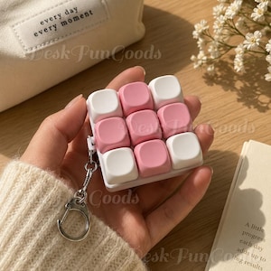 Puede incluir: Una funda protectora cuadrada para auriculares inalámbricos con una rejilla táctil de nueve botones elevados y redondeados en colores blanco y rosa suave alternados. La funda incluye un llavero de metal plateado en el lateral.