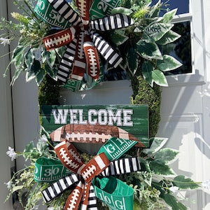 May include: A decorative wreath featuring a moss-covered base, green leaves, and white flowers. It includes two large bows made of ribbons with football, yard line, and black-and-white striped patterns. A central green sign reads WELCOME.