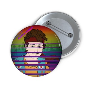 May include: A round pin-back button featuring a colourful illustration of a person with brown hair wearing a red headband and a patterned vest. The text Welcome to SANTA CARLA is printed in purple. The design includes horizontal stripes.