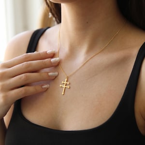 Puede incluir: Un collar de tono dorado con un colgante en forma de cruz de doble barra, también conocida como cruz de Lorena. El colgante cuelga de una cadena fina y delicada de tono dorado. El diseño es sencillo, con líneas limpias y acabado pulido.