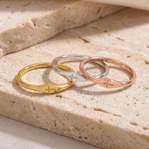 Puede incluir: Tres anillos finos de metal en tonos oro, plata y oro rosa. Cada anillo presenta una placa plana centrada grabada con un pequeño símbolo de cruz. Las bandas son lisas y circulares, mostrando un acabado metálico pulido en cada pieza.