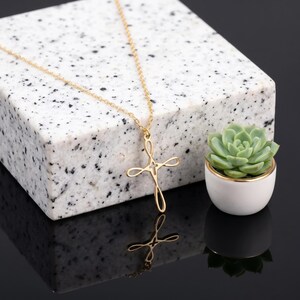 May include: A gold-toned necklace featuring a delicate, open-work cross pendant with flowing, curved lines. The pendant hangs from a thin, matching gold-toned chain, creating a refined and simple aesthetic that highlights the metallic finish.