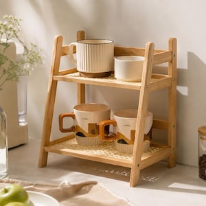 May include: A two-tier wooden shelf with a woven cane base on each level. The top shelf holds a tall, vertically ribbed cream mug and a small plain white bowl. The bottom shelf holds two cream mugs featuring a brown and black landscape graphic.