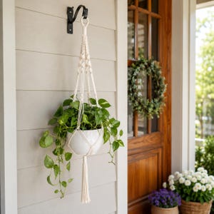 Op de afbeelding: Een crèmekleurige macramé plantenhanger van geknoopt koord, waaraan een gladde witte keramische pot hangt met een weelderige groene pothosplant. De hanger heeft een decoratieve kwast aan de onderkant en hangt aan een zwarte metalen muurhaak.
