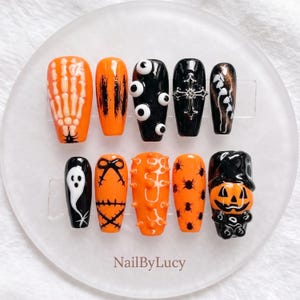 Könnte beinhalten: Ein Set aus zehn künstlichen Fingernägeln mit Halloween-Motiven in Orange, Schwarz und Weiß. Die Designs umfassen eine Skeletthand, Augäpfel, ein silbernes Kreuz, ein Gespenst, Spinnen und einen Kürbis. Der Text NailByLucy ist unten aufgedruckt.