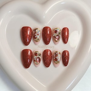 Op de afbeelding: Een set van tien kunstnagels met een dieprode glitterafwerking en accentnagels op een lichtroze basis, versierd met goudomrande rode en iriserende strass-steentjes. De tekst NailByLucy staat in het midden van de display gedrukt.