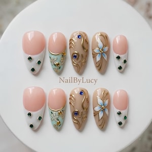 Könnte beinhalten: Ein Set aus zehn künstlichen Fingernägeln mit dem Text NailByLucy in der Mitte. Die Designs umfassen weiße French-Tips mit grünen Strasssteinen, hellgrüne Blumenmuster mit Goldakzenten, beige Schnörkel und weiße Blumenmalereien.