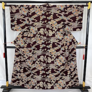 May include: A dark purple kimono featuring a repeating pattern of colorful floral clusters and swirling wave-like lines in shades of blue, orange, yellow, and green. The garment has wide sleeves and is displayed on a black wooden stand with red tassels.