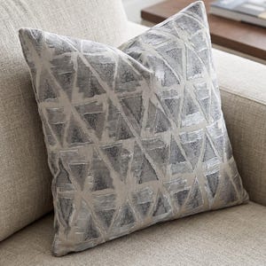 Può includere: Un cuscino quadrato con un motivo geometrico ripetuto di triangoli grigio argento su sfondo beige chiaro. Le forme triangolari presentano una lucentezza metallica testurizzata che contrasta con la base in tessuto liscio di colore neutro.