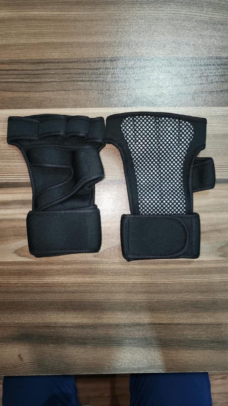May include: A pair of black fingerless hand supports featuring a wrap-around wrist strap with a hook-and-loop closure. One support shows the palm side with a textured grip, while the other displays the back side with a breathable mesh panel.
