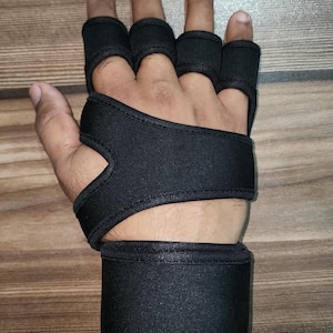May include: A black fingerless hand support brace featuring individual loops for four fingers, a wide palm strap, and a thick adjustable wrist wrap. The brace is constructed from a flexible, solid black fabric material designed to provide compression.