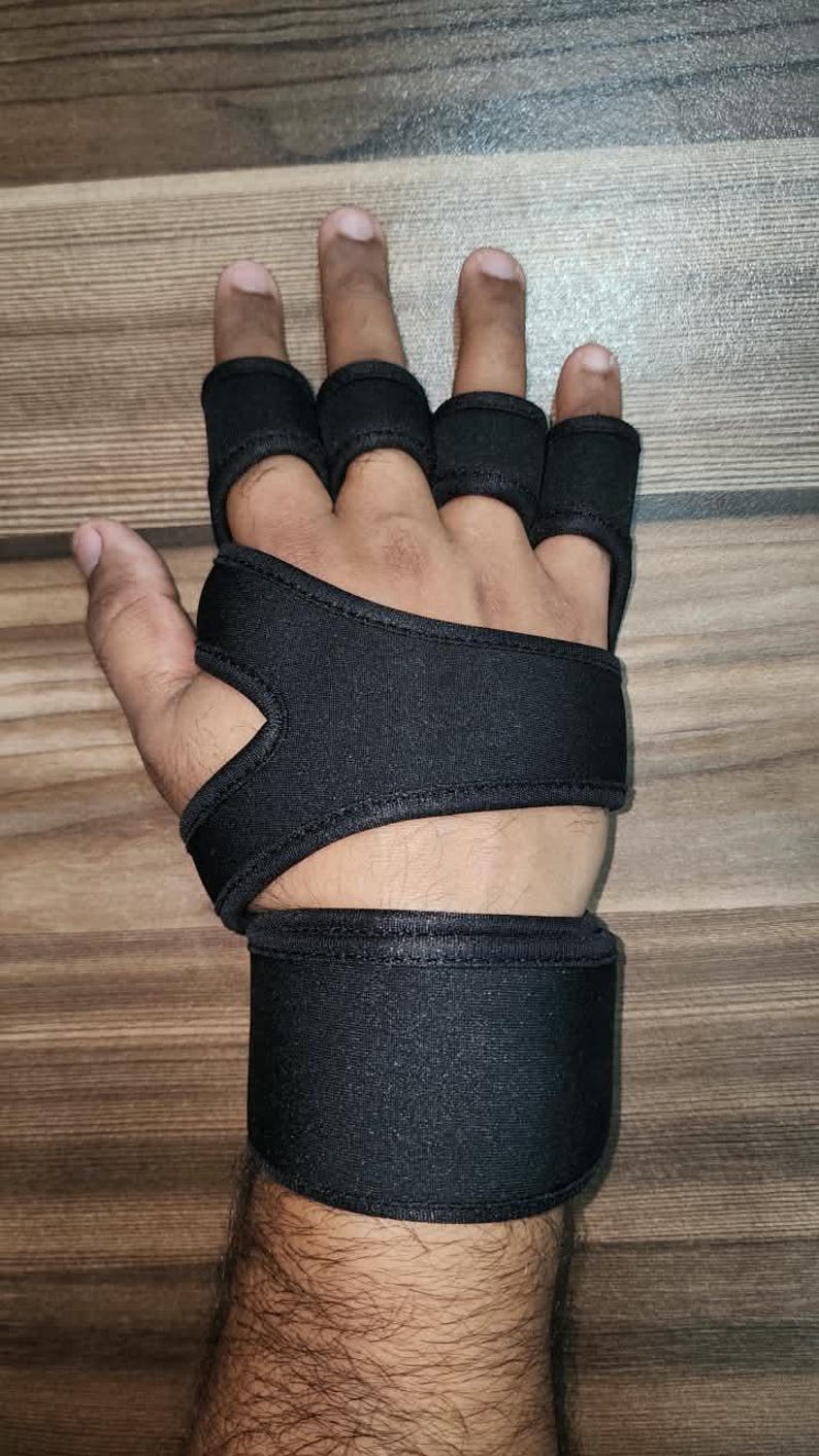 May include: A black fingerless hand support brace featuring individual loops for four fingers, a wide palm strap, and a thick adjustable wrist wrap. The brace is constructed from a flexible, solid black fabric material designed to provide compression.