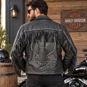 May include: A black leather jacket featuring long fringe detailing across the back and sleeves, adorned with silver metal studs and an embossed eagle graphic above the Harley-Davidson logo on the back panel, worn with dark denim jeans.