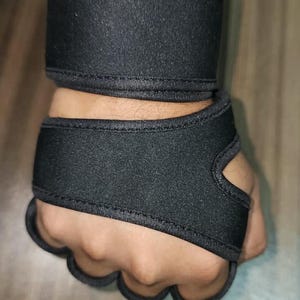 May include: A black wrist and hand support brace featuring a wide band that wraps around the wrist and a secondary section that extends over the back of the hand with individual finger loops to secure the wrap in place around the palm area.