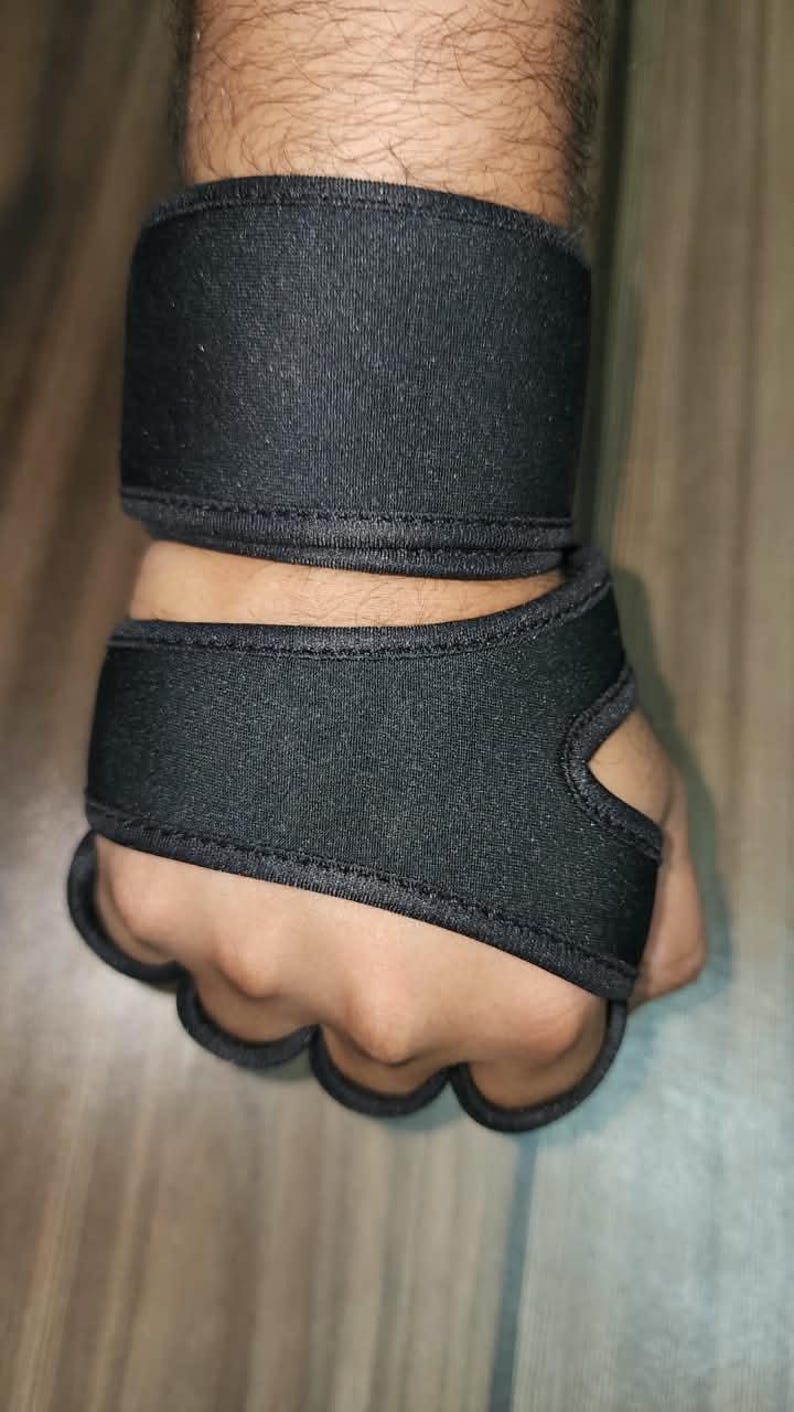 May include: A black wrist and hand support brace featuring a wide band that wraps around the wrist and a secondary section that extends over the back of the hand with individual finger loops to secure the wrap in place around the palm area.