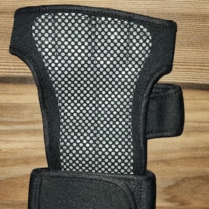 May include: A black wrist support brace featuring a central panel with a white perforated mesh pattern. The brace includes a wide adjustable strap at the base and a smaller side strap, both secured with hook-and-loop fasteners for a snug fit.