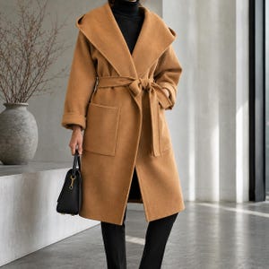 May include: A camel-coloured hooded coat featuring a wide lapel, long sleeves with folded cuffs, and a matching tie belt at the waist. The coat includes two large patch pockets on the front and is worn over a black turtleneck and black trousers.