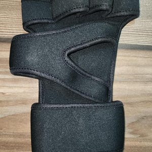 May include: A black fingerless hand support brace made of neoprene-like material. It features a wide wrist strap with a hook-and-loop closure, a thumb opening, and individual finger loops across the top section to provide stability and compression.