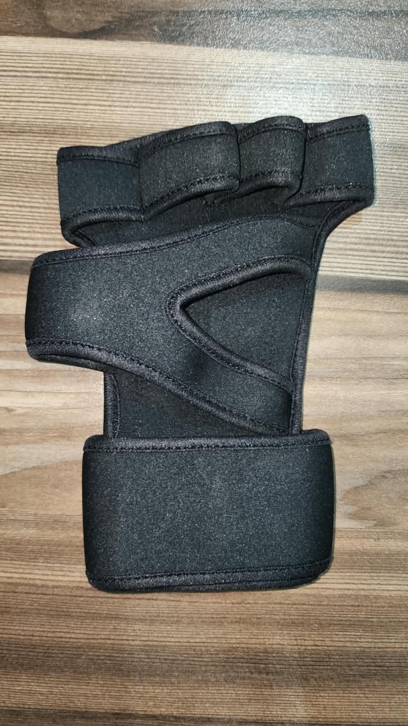 May include: A black fingerless hand support brace made of neoprene-like material. It features a wide wrist strap with a hook-and-loop closure, a thumb opening, and individual finger loops across the top section to provide stability and compression.