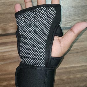 May include: A black fingerless glove with a grey mesh palm pad featuring a circular dot pattern. The glove includes a wide black wrist strap with a hook-and-loop closure, designed to provide support and grip for various physical activities.