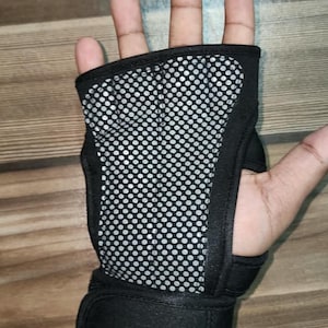 May include: A black fingerless hand brace featuring a central panel with a silver and black circular mesh pattern. The brace includes a wide adjustable strap at the wrist with a hook-and-loop closure, designed to provide support to the palm and wrist.