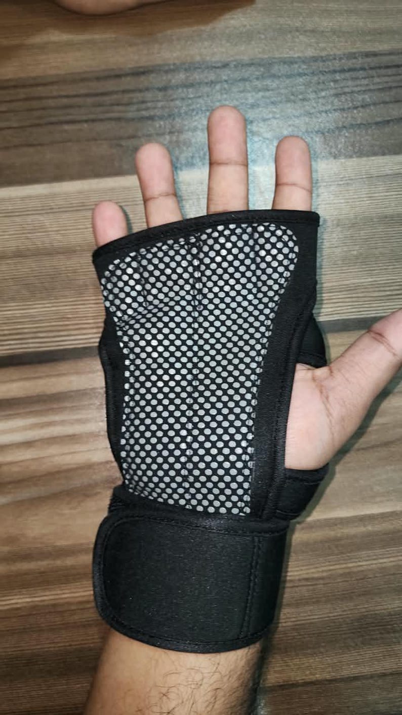 May include: A black fingerless hand brace featuring a central panel with a silver and black circular mesh pattern. The brace includes a wide adjustable strap at the wrist with a hook-and-loop closure, designed to provide support to the palm and wrist.