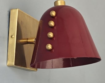 May include: A wall sconce featuring a deep burgundy, bell-shaped shade with a glossy finish. The fixture includes a rectangular brushed brass wall plate and a matching brass arm. Four small, spherical brass studs are aligned vertically on the shade.