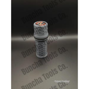Op de afbeelding: Een cilindrische grijze opbergcontainer met een gestructureerd zeshoekig patroon aan de buitenkant. Het bovendeksel bevat een rond logo met de tekst STASH TUBE en een gele bliksemschicht. De container heeft een duidelijke horizontale naad.