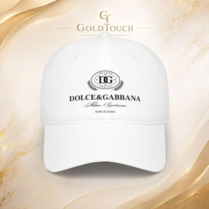 May include: A white baseball cap featuring a black embroidered logo on the front. The logo includes the text DG Milano Italia, Dolce & Gabbana, Italian Sportswear, and Since:0084. The cap has a structured crown and a curved brim.