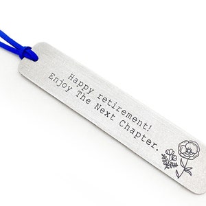 May include: A silver metal bookmark with the text "Happy retirement! Enjoy The Next Chapter." and a floral design. It has a blue ribbon attached at the top for easy placement in a book.
