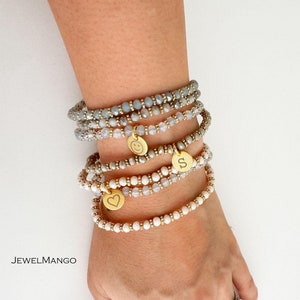 Puede incluir: Conjunto de pulseras de cuentas en tonos grises, blancos y dorados. Cada pulsera presenta pequeñas cuentas y detalles dorados. Algunas pulseras tienen colgantes dorados con una carita sonriente, un corazón y la letra "S". La marca "JEWELMANGO" es visible.