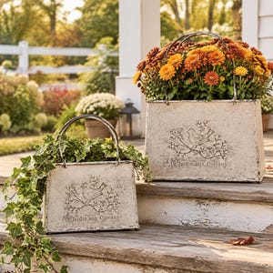 May include: Two off-white metal planters with arched handles, each featuring an embossed design of a bird perched on a branch and the text Le Jardin aux Oiseaux. The larger planter holds orange chrysanthemums, while the smaller holds ivy.