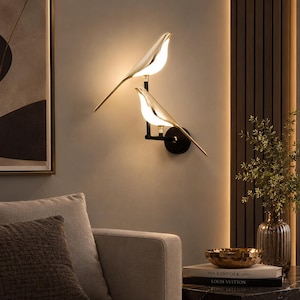 Könnte beinhalten: Eine Wandleuchte mit zwei vogelförmigen Lampen, die goldene Metallkörper und leuchtend weiße Unterseiten haben. Die Vögel sind an einem schwarzen Metallrahmen befestigt, der mit einer runden, schwarzen Wandhalterung verbunden ist.