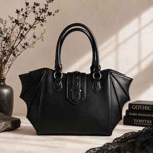 Könnte beinhalten: Eine schwarze Handtasche mit seitlichen Einsätzen in Form von Fledermausflügeln, zwei Tragegriffen und einem mittigen vertikalen Riemenverschluss mit Metallnieten. Die Tasche hat eine glatte Oberfläche und eine strukturierte Form.