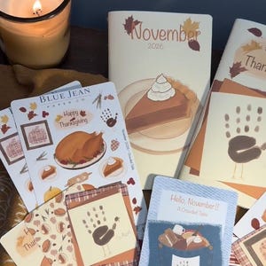 May include: A collection of Thanksgiving-themed paper stickers and cards from Blue Jean Paper Co. featuring illustrations of a roasted turkey, pumpkin pie, and handprint turkeys, alongside a lit candle and a November 2026 calendar.
