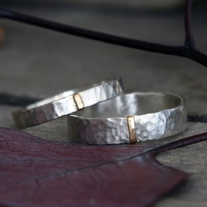 Pode incluir: Dois anéis de prata com textura martelada e uma única barra vertical dourada incrustada em cada banda. Os anéis estão posicionados perto de uma folha roxa escura, exibindo o acabamento metálico e o contraste entre os elementos prateados e dourados.